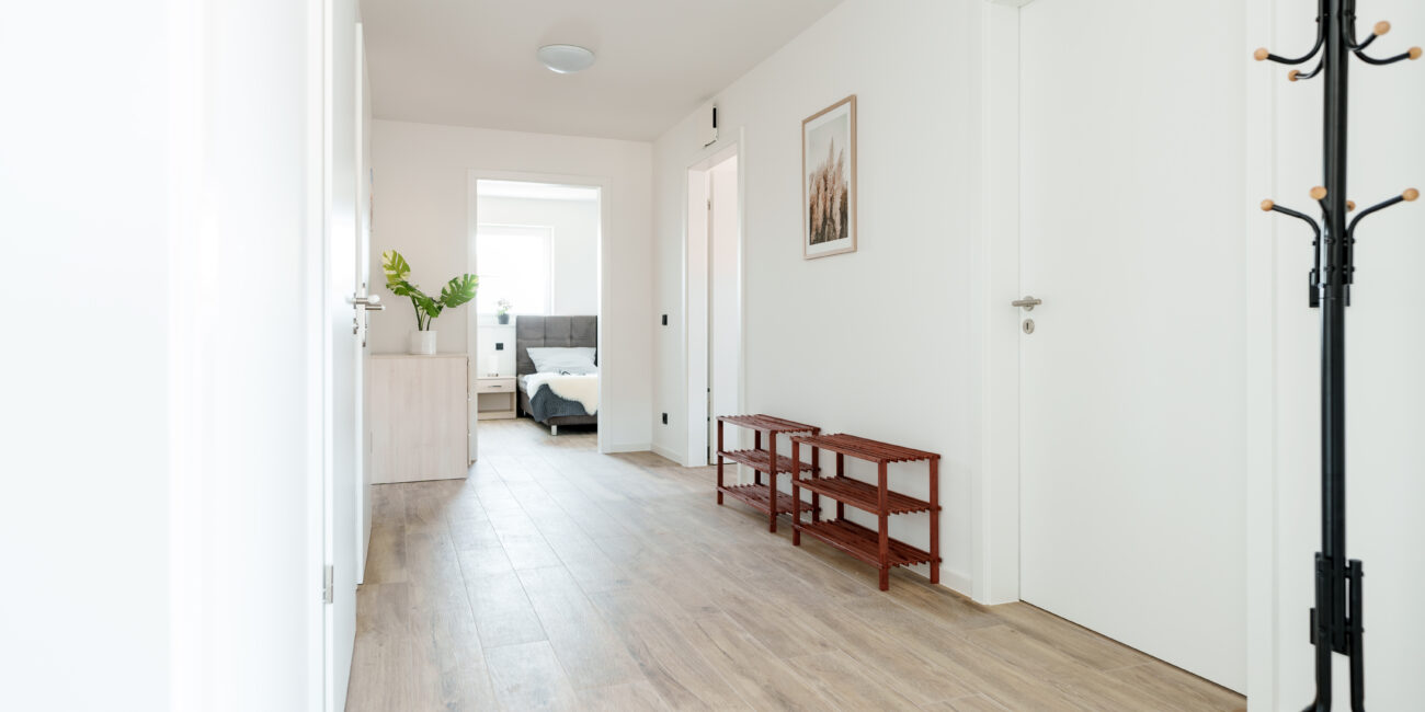 Heller Flur mit Holzoptik-Boden, weißen Türen, Schuhregalen, Garderobenständer und Blick in ein Schlafzimmer mit grauem Polsterbett.