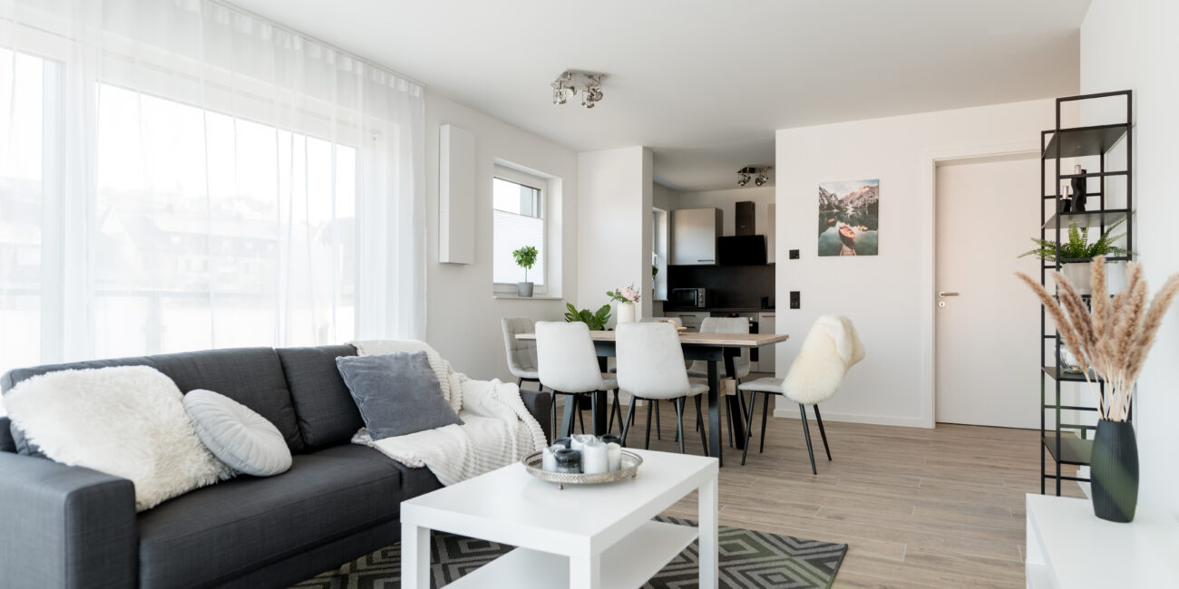 Offener Wohn- und Essbereich mit grauem Sofa, Esstisch mit sechs Stühlen, weißem Couchtisch, Teppich und Blick zur Küche im Hintergrund.