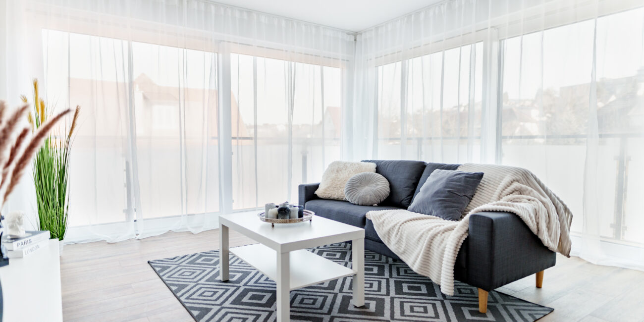 Helles Wohnzimmer mit Ecksofa, weißen Vorhängen, geometrischem Teppich, Couchtisch und großer Fensterfront mit Blick ins Freie.