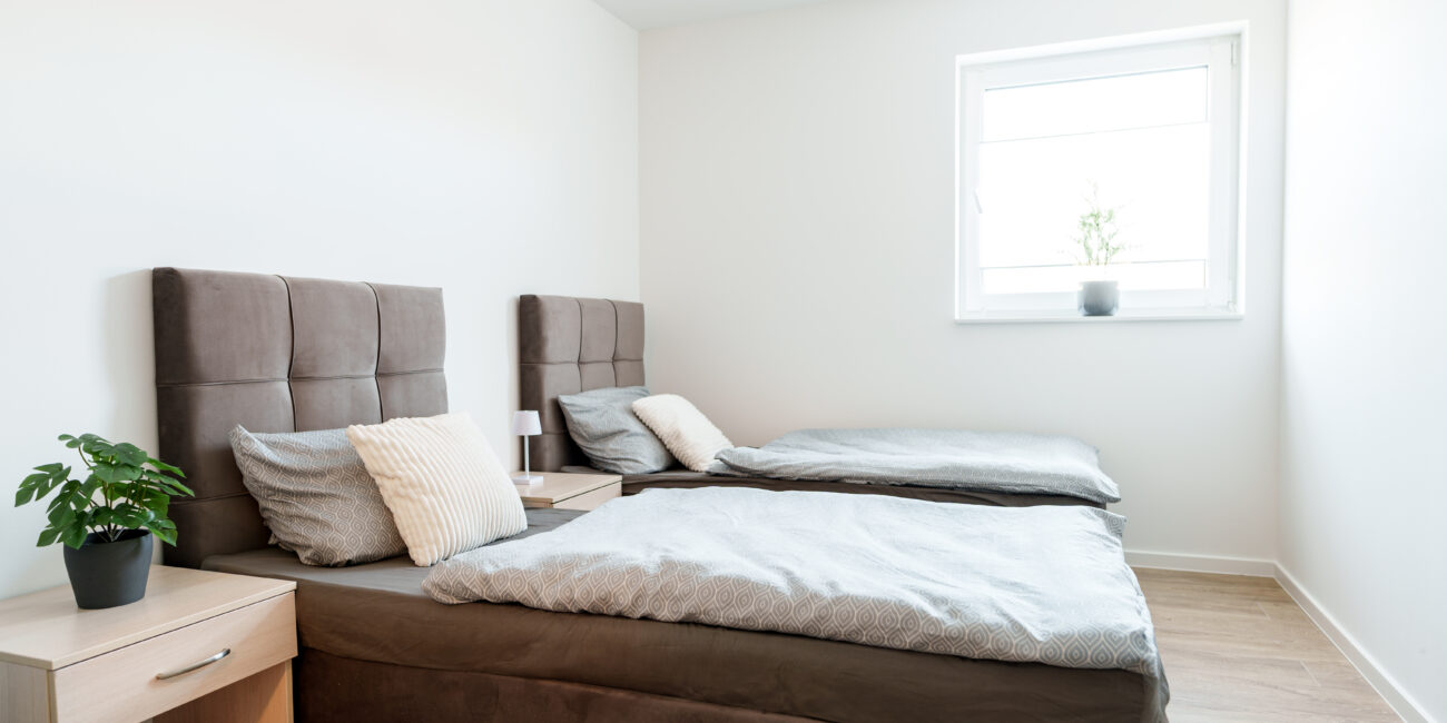 Helles Schlafzimmer mit zwei Einzelbetten, grauen Polsterkopfteilen, Holzboden, Nachttischen und einem Fenster mit Topfpflanze.