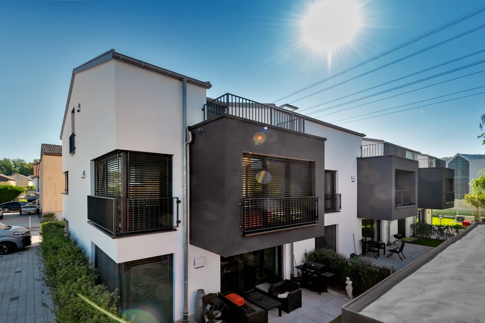Modernes Wohngebäude mit schwarzen Balkonen und einem Dachgarten, sonnig mit klar blauem Himmel und Blick auf die Terrasse mit Gartenmöbeln.
