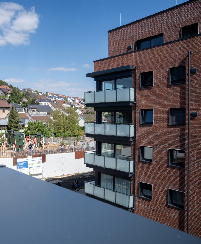 Backsteinwohnhaus mit Glasbalkonen, Blick auf einen Spielplatz und Wohnsiedlung im Hintergrund bei klarem Himmel.